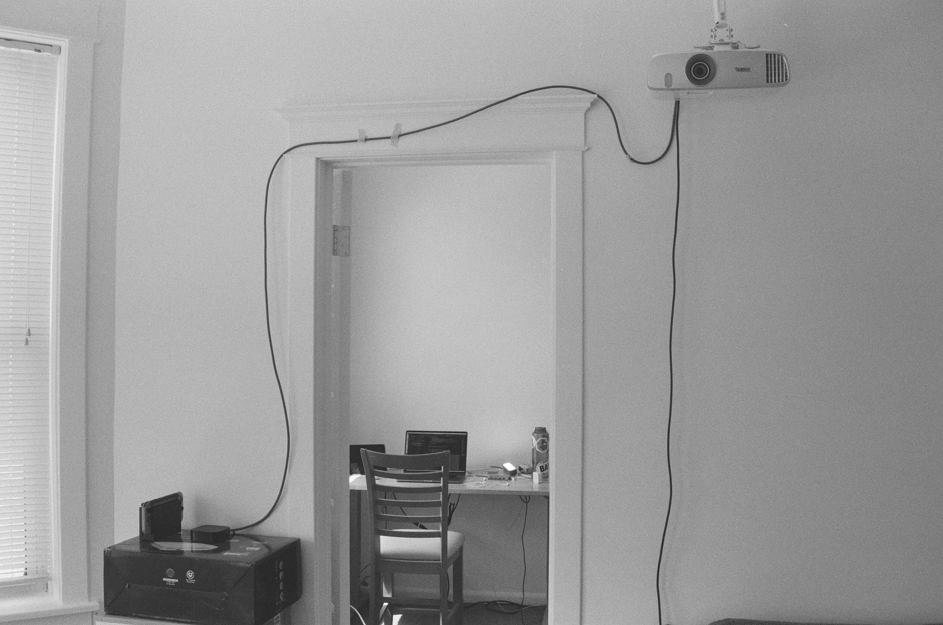 A picture of a doorway to a desk with a laptop on it. In front of the doorway, a cord is run sloppily from the bottom left across the top to a ceiling mounted projector.